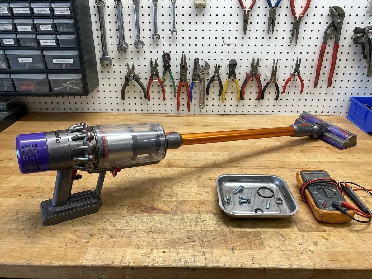 Dyson cordless stick vacuum on a workbench next to a parts tray and a multimeter