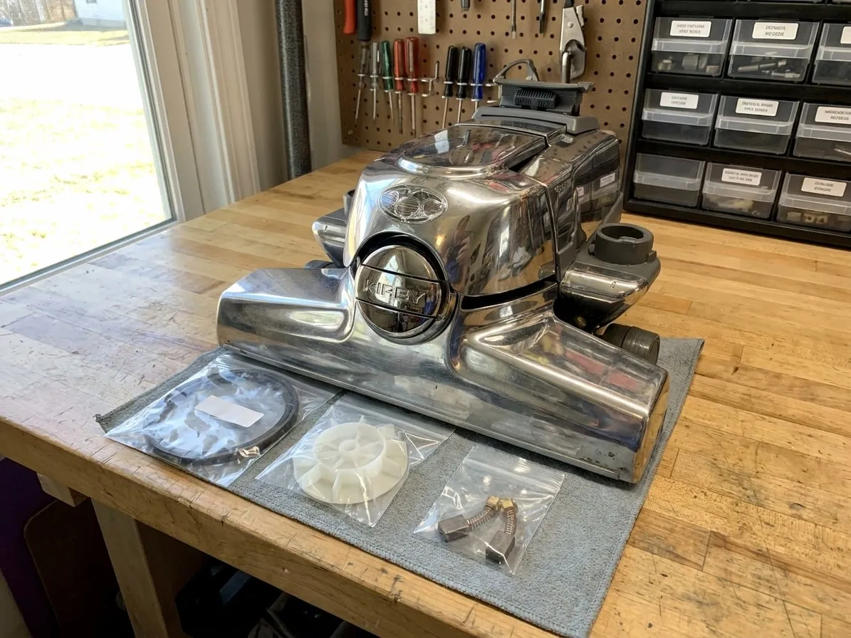 Polished Kirby motor housing and headlight beside new belts, a fan impeller and motor brushes